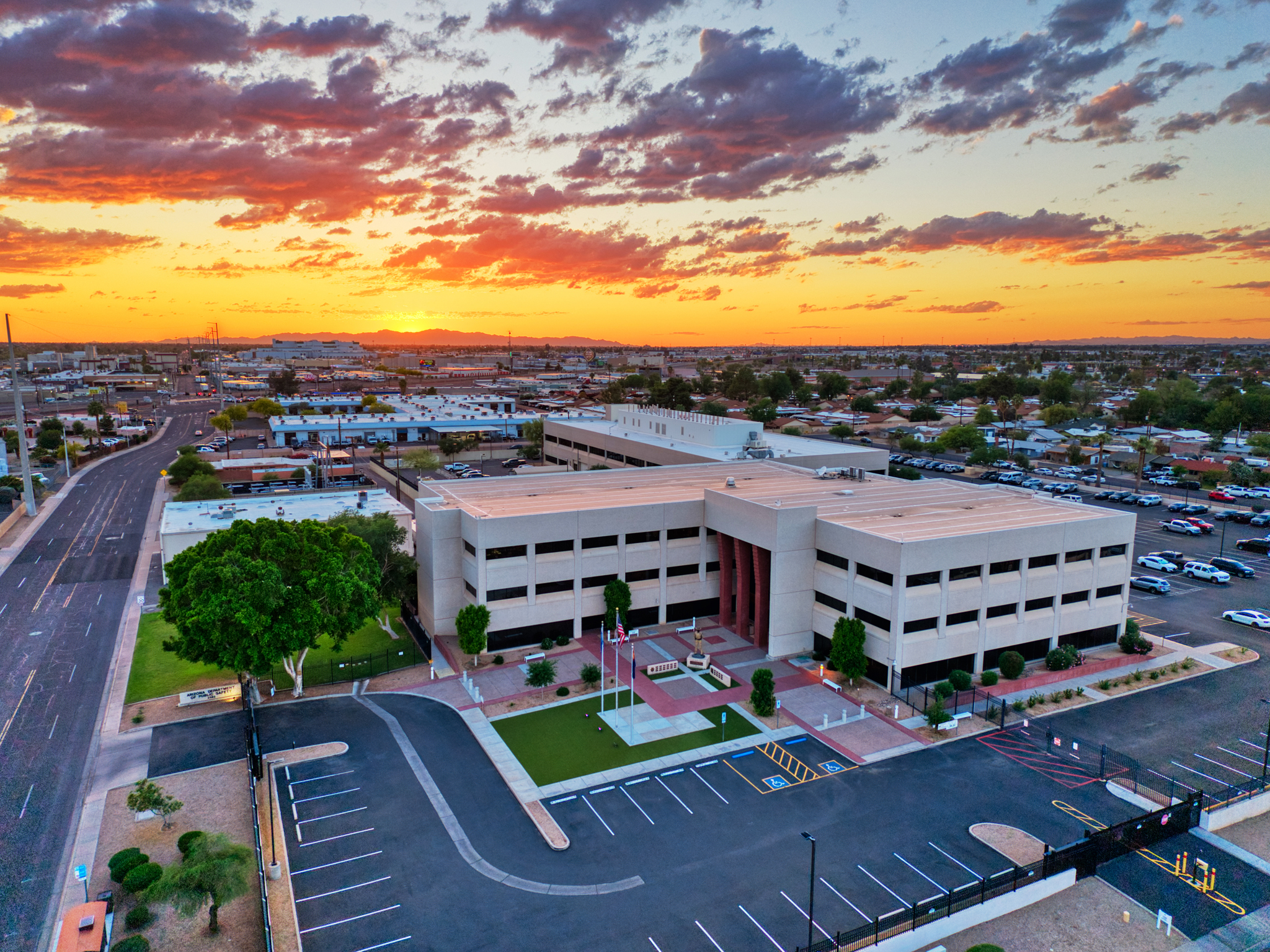 Wide aerial view of the Arizona Department of Public Safety (DPS) Headquarters in Phoenix at sunset, featuring the multi-story office building, parking lot, and a vibrant orange sky.
