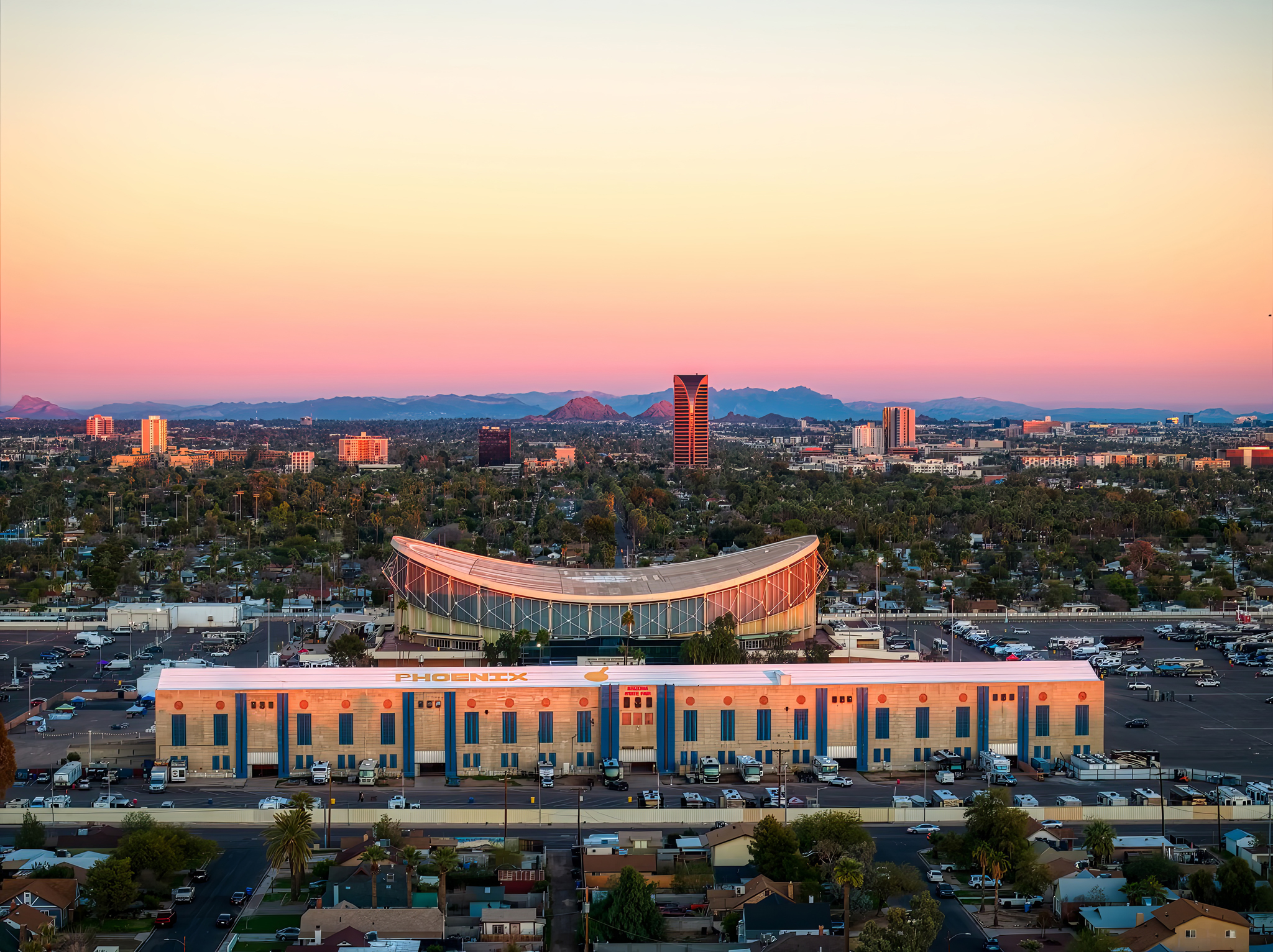 Wide aerial view of the Arizona State Fairgrounds in Phoenix at sunset, featuring the Veterans Memorial Coliseum and the city skyline under a warm orange and pink sky.