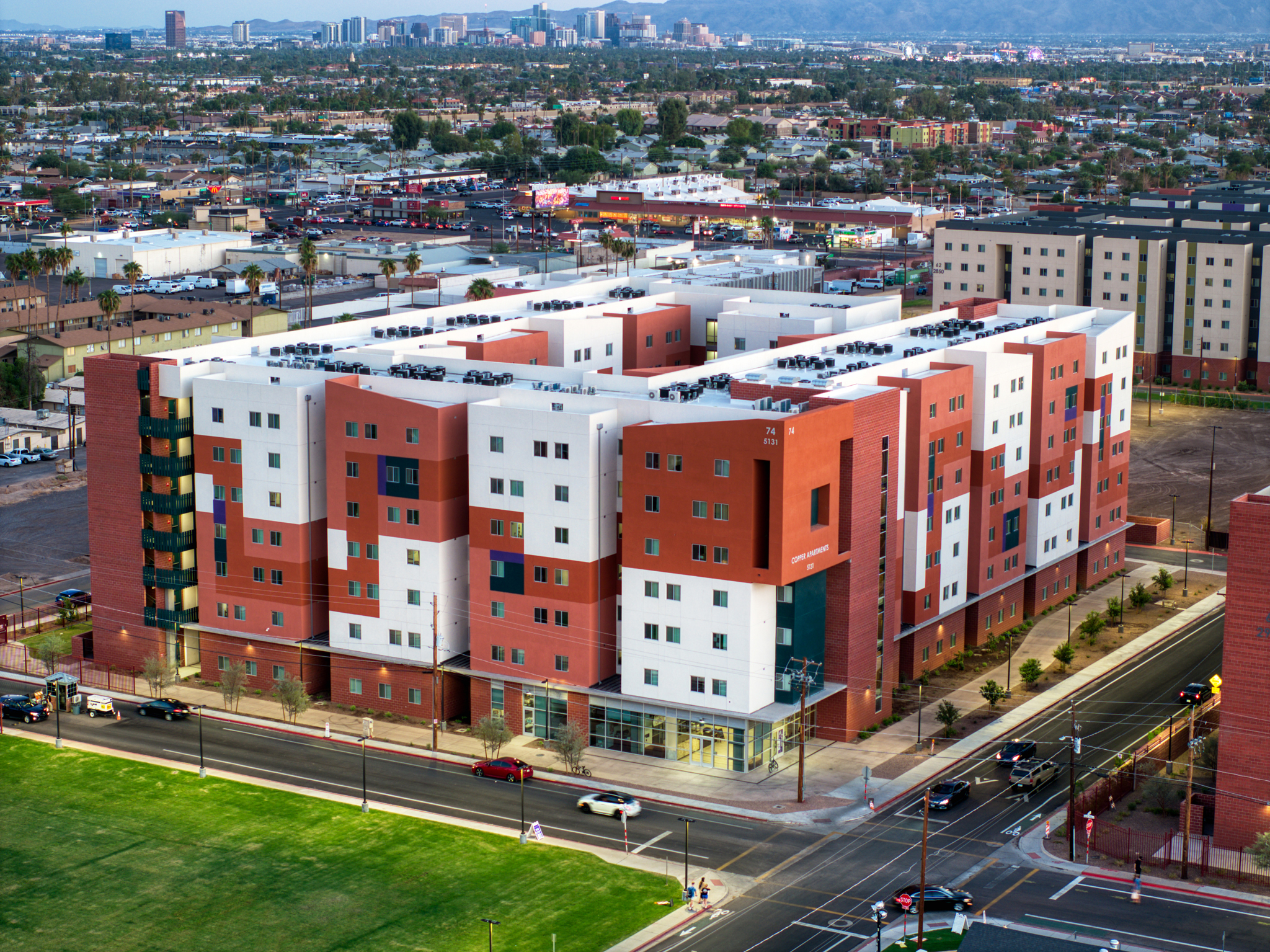A high-angle aerial drone shot of the GCU Copper Apartments in Phoenix, Arizona, showing the modern multi-story residential building with its distinct orange and white facade, adjacent roads, and the city skyline in the distance.