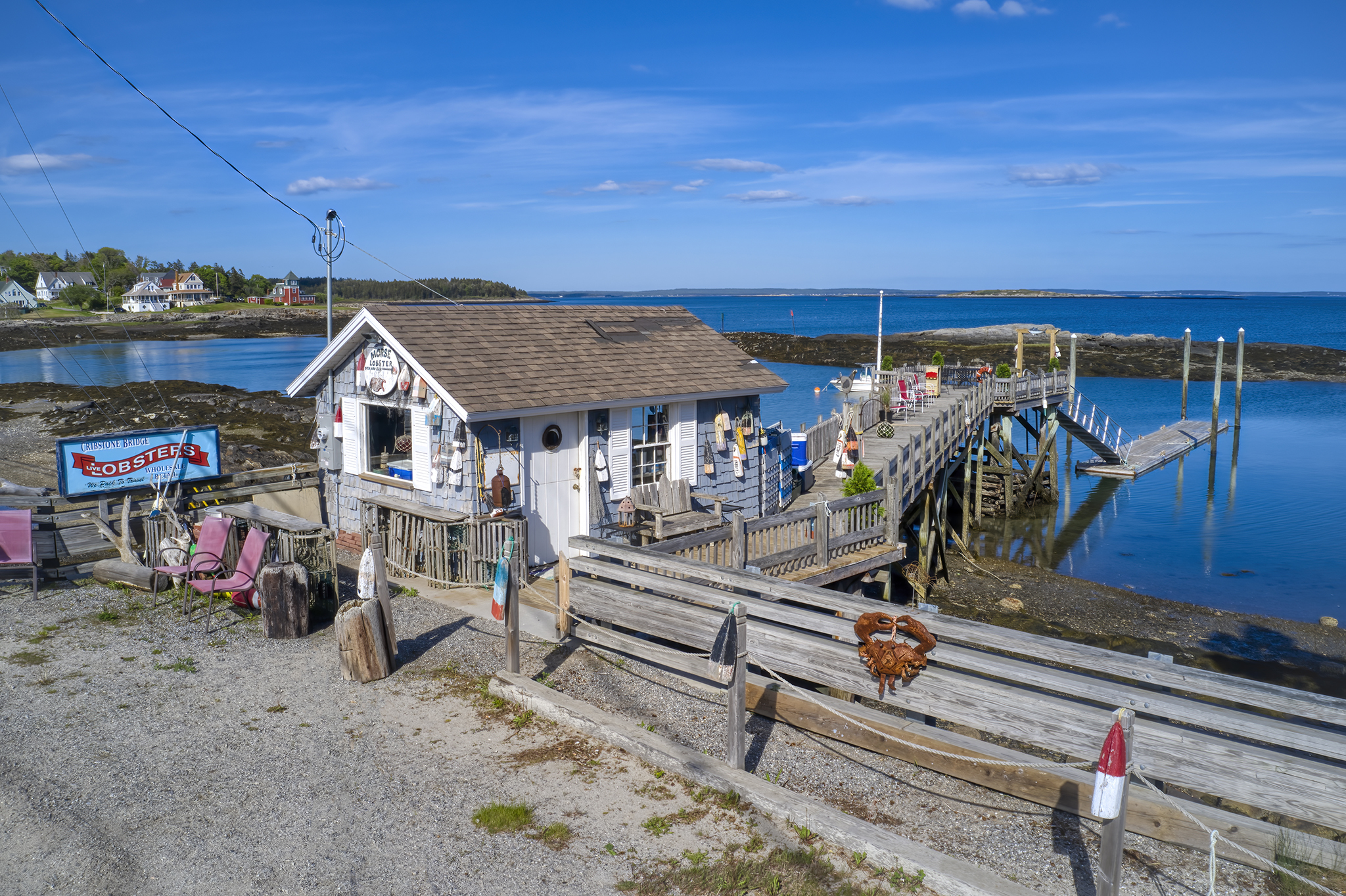 An aerial drone perspective of a traditional weathered Maine lobster shack and a wooden pier extending over the water on a clear, sunny day. A red lobster trap prop is prominent in the foreground.