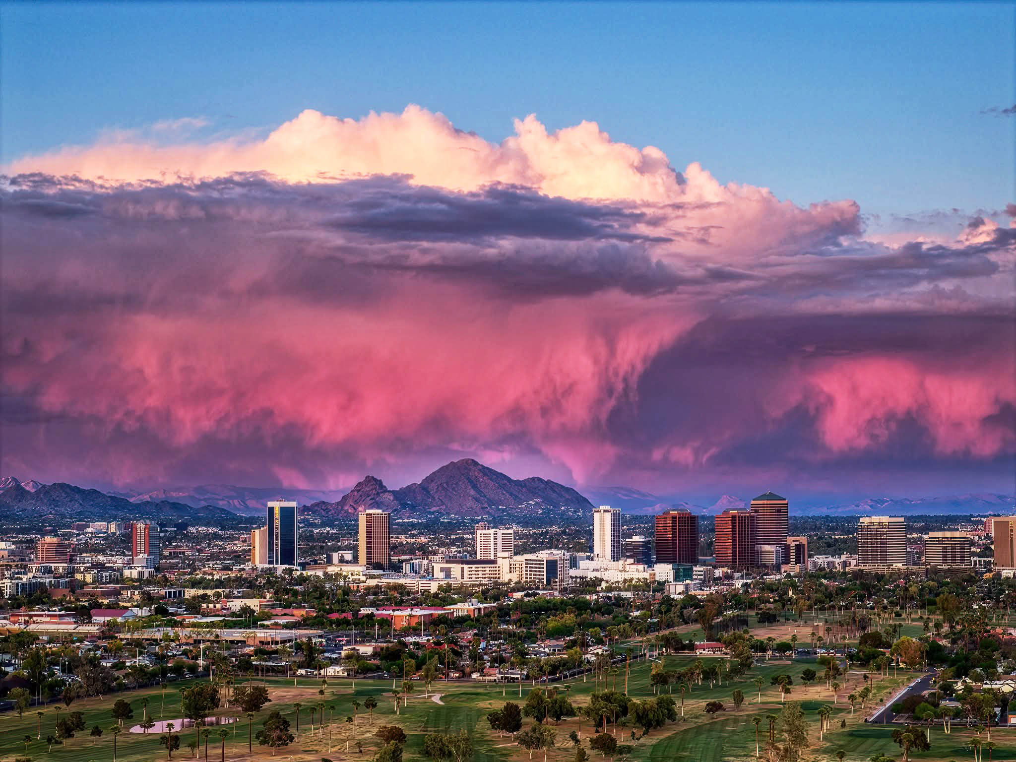 Wide aerial view of downtown Phoenix skyscrapers at sunset under a massive bank of glowing pink and purple monsoon clouds.